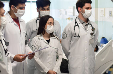 Grupo de estudantes de Medicina da Unoeste em visita hospitalar, usando jaleco branco, estetoscópio e máscara, acompanhando uma atividade prática ao lado de leitos hospitalares como parte da formação ética e clínica.