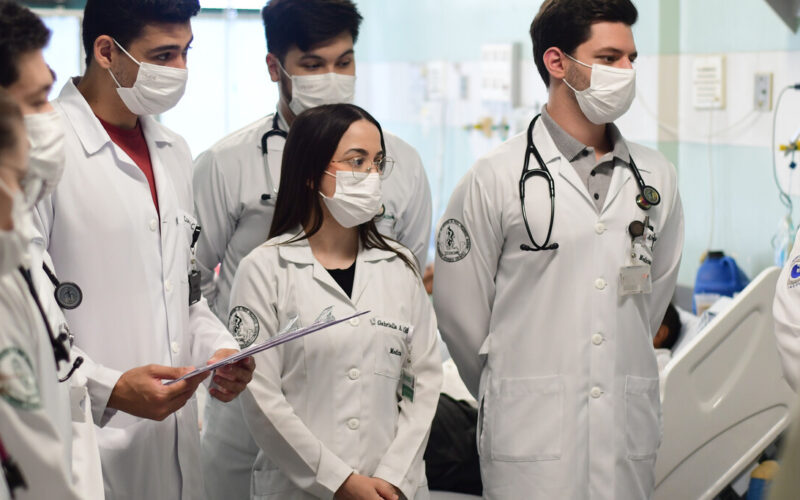 Grupo de estudantes de Medicina da Unoeste em visita hospitalar, usando jaleco branco, estetoscópio e máscara, acompanhando uma atividade prática ao lado de leitos hospitalares como parte da formação ética e clínica.