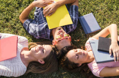 Três jovens deitados sobre a grama, em formato de círculo, segurando livros e cadernos coloridos, representando o universo da leitura entre adolescentes e o hábito de ler livros Young Adult antes da faculdade.