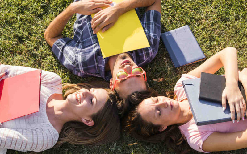 Três jovens deitados sobre a grama, em formato de círculo, segurando livros e cadernos coloridos, representando o universo da leitura entre adolescentes e o hábito de ler livros Young Adult antes da faculdade.