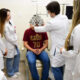 Estudantes de Medicina da Unoeste, todos de jaleco branco, acompanham demonstração prática de exame oftalmológico realizada por professor em paciente, que está sentado em cadeira de atendimento e usando um equipamento de refração ocular em ambiente clínico. A cena representa o aprendizado técnico durante a formação médica