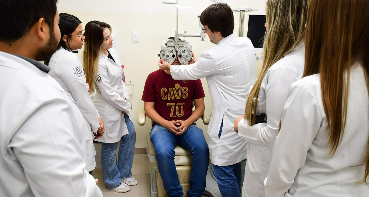 Estudantes de Medicina da Unoeste, todos de jaleco branco, acompanham demonstração prática de exame oftalmológico realizada por professor em paciente, que está sentado em cadeira de atendimento e usando um equipamento de refração ocular em ambiente clínico. A cena representa o aprendizado técnico durante a formação médica