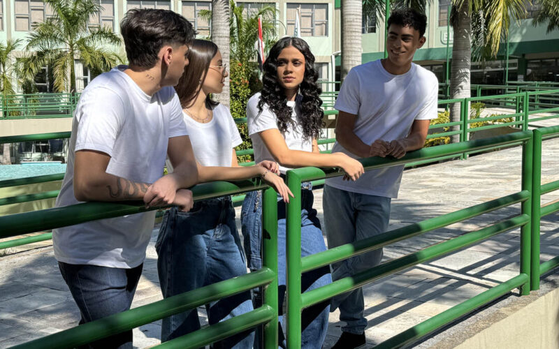 Quatro estudantes universitários conversam ao ar livre no campus da Unoeste, cercados por palmeiras e edifícios modernos, vestindo camisetas brancas e calças jeans, em um ambiente descontraído e acolhedor.