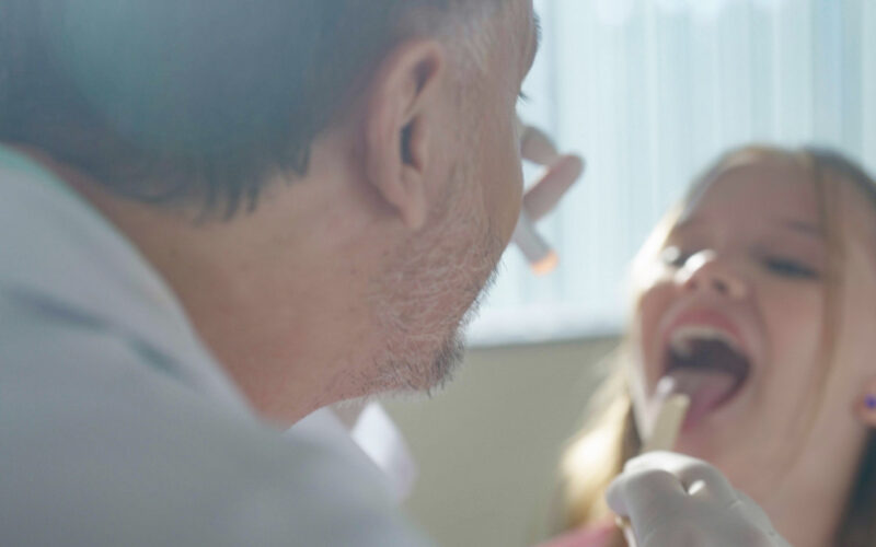 Médico examina a garganta de uma criança durante atendimento clínico, utilizando abaixador de língua e lanterna, em consultório com luz natural.