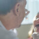 Médico examina a garganta de uma criança durante atendimento clínico, utilizando abaixador de língua e lanterna, em consultório com luz natural.