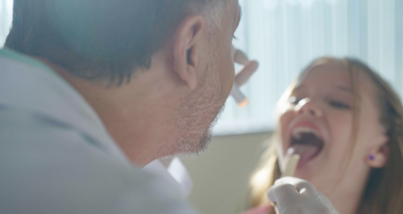 Médico examina a garganta de uma criança durante atendimento clínico, utilizando abaixador de língua e lanterna, em consultório com luz natural.