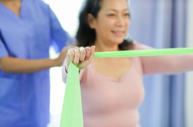 Mulher realizando exercício com faixa elástica durante sessão de fisioterapia, acompanhada por profissional da área da saúde. Técnica utilizada na Reeducação Postural Global (RPG).