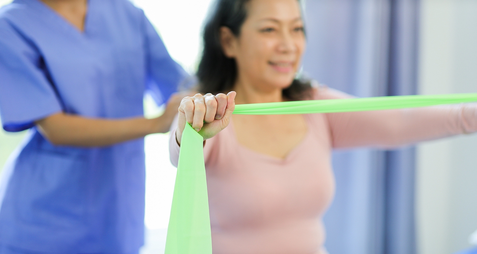 Mulher realizando exercício com faixa elástica durante sessão de fisioterapia, acompanhada por profissional da área da saúde. Técnica utilizada na Reeducação Postural Global (RPG).