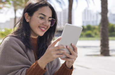 Jovem sorridente usando um leitor digital Kindle ao ar livre, com óculos de grau e cabelo solto, sentada em ambiente urbano com palmeiras ao fundo.
