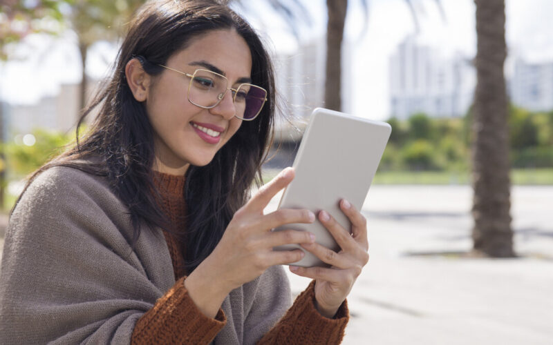 Jovem sorridente usando um leitor digital Kindle ao ar livre, com óculos de grau e cabelo solto, sentada em ambiente urbano com palmeiras ao fundo.