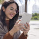 Jovem sorridente usando um leitor digital Kindle ao ar livre, com óculos de grau e cabelo solto, sentada em ambiente urbano com palmeiras ao fundo.