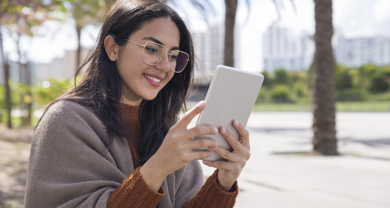 Jovem sorridente usando um leitor digital Kindle ao ar livre, com óculos de grau e cabelo solto, sentada em ambiente urbano com palmeiras ao fundo.