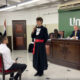 Estudantes de Direito da Unoeste participam de um júri simulado, com um aluno vestido de toga interrogando uma testemunha sentada, enquanto professores e avaliadores acompanham a atividade no plenário.