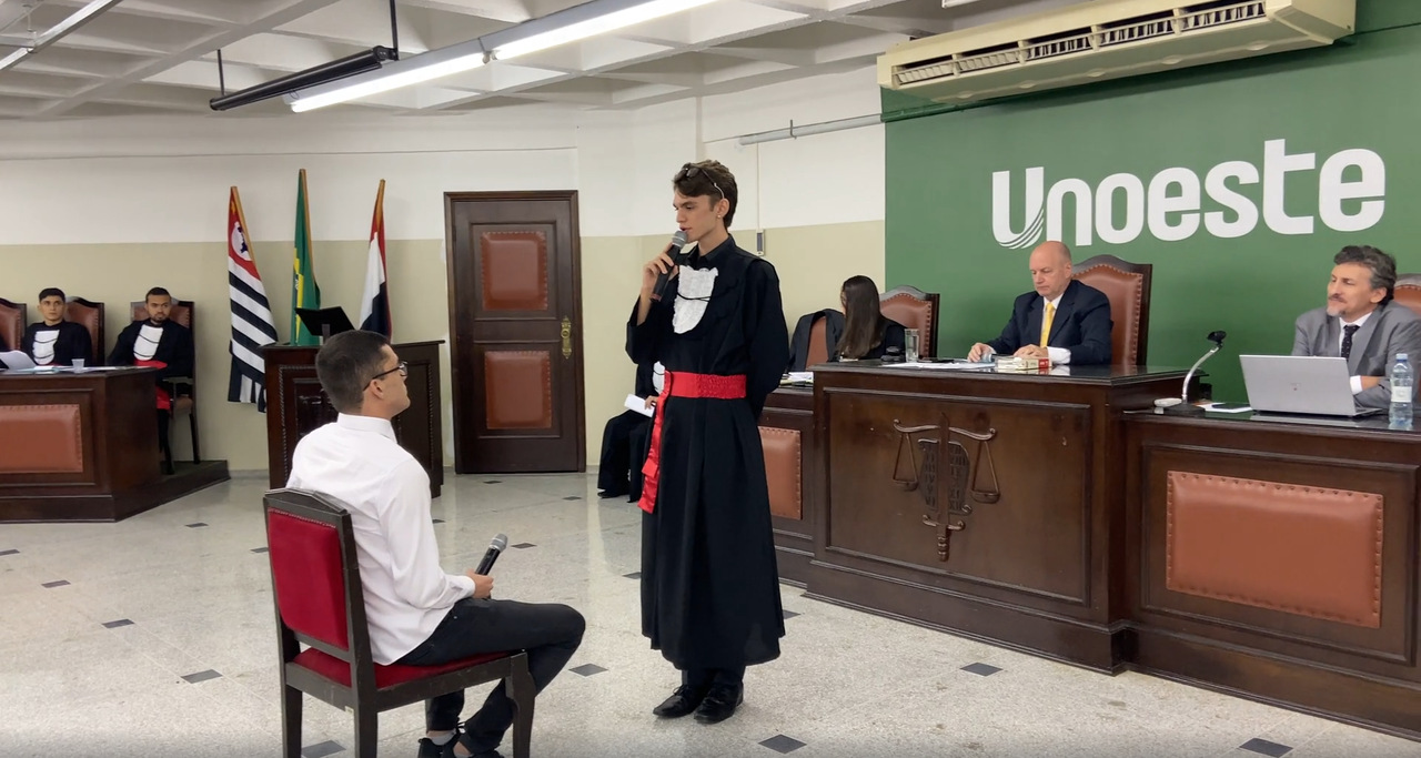 Estudantes de Direito da Unoeste participam de um júri simulado, com um aluno vestido de toga interrogando uma testemunha sentada, enquanto professores e avaliadores acompanham a atividade no plenário.