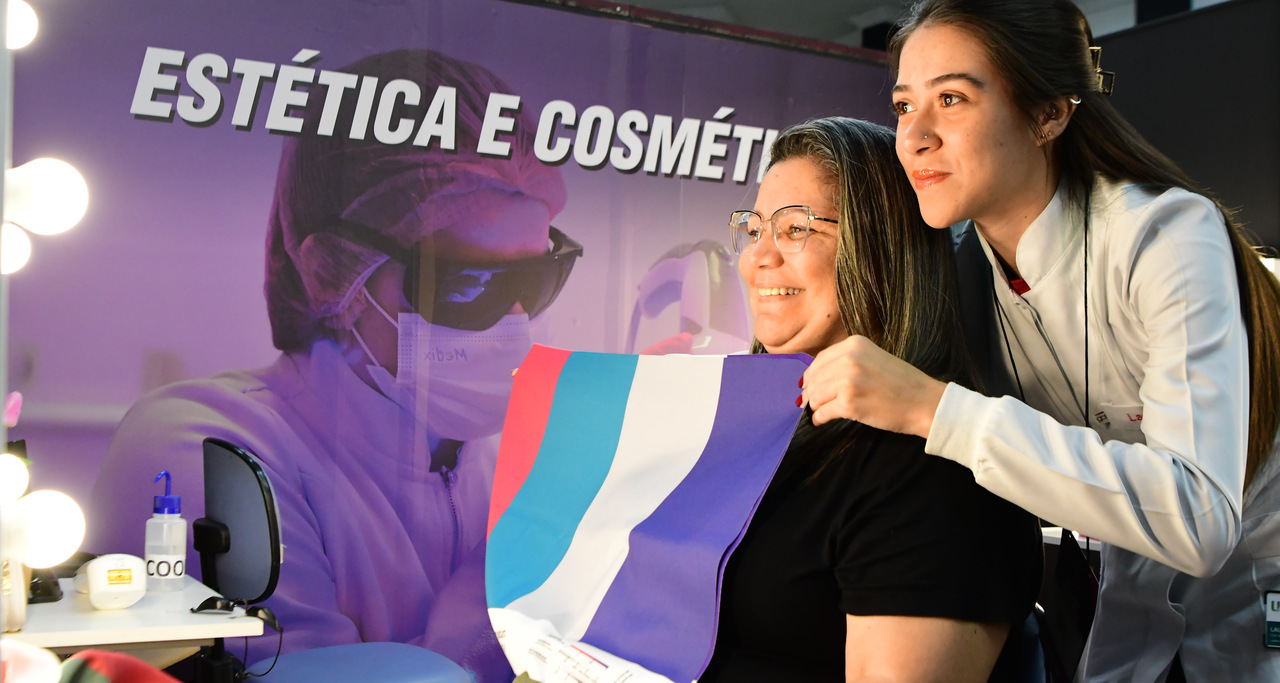 Aluna do curso de Estética e Cosmética da Unoeste orienta visitante durante feira de profissões, mostrando possibilidades da carreira na área da beleza e bem-estar.