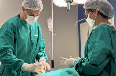 Estudante de Medicina da Unoeste realiza procedimento prático supervisionado em ambiente hospitalar, vestindo roupas cirúrgicas e máscara, durante atividade de treinamento.