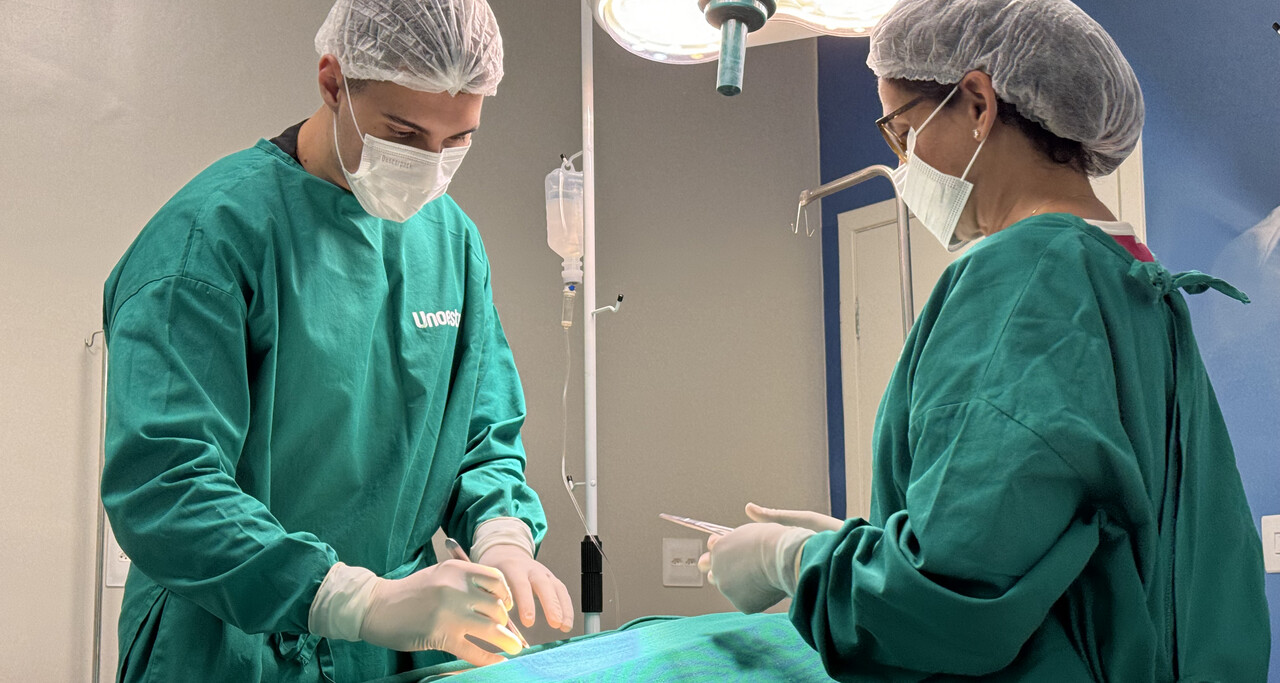 Estudante de Medicina da Unoeste realiza procedimento prático supervisionado em ambiente hospitalar, vestindo roupas cirúrgicas e máscara, durante atividade de treinamento.