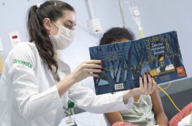 Estudante da Unoeste com jaleco branco lê um livro infantil para uma criança em ambiente hospitalar durante atividade do Projeto Leia no Leito, demonstrando empatia e atuação como voluntária no cuidado humanizado.