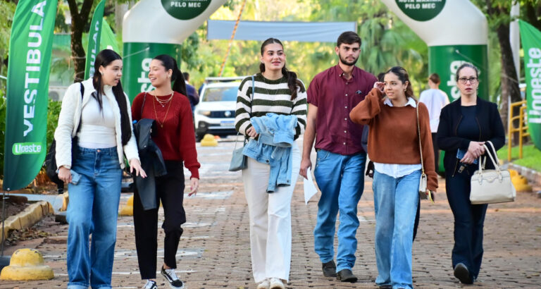 Estudantes caminham juntos pelo campus da Unoeste, em área externa sinalizada com bandeiras da universidade.