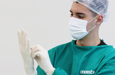 Estudante de Medicina da Unoeste vestindo avental cirúrgico verde, máscara e touca descartável, colocando luvas estéreis antes de iniciar atividade prática em ambiente hospitalar.