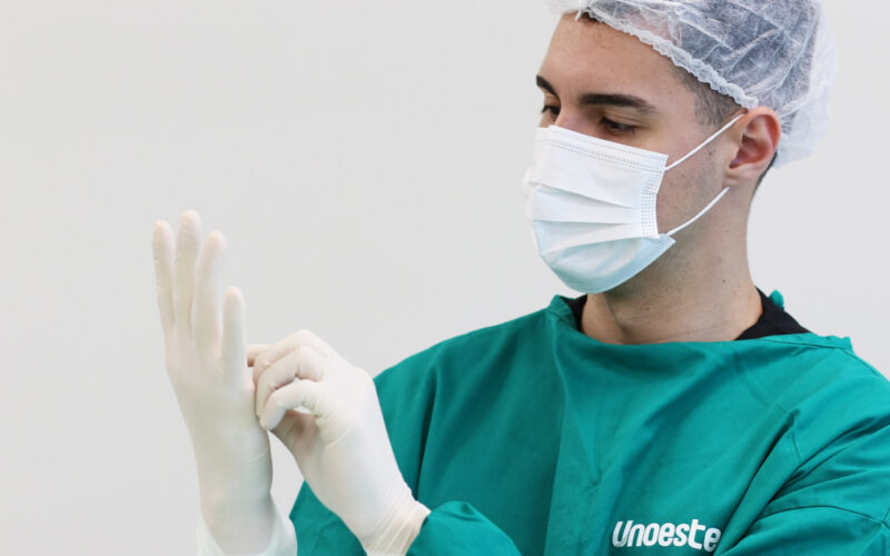 Estudante de Medicina da Unoeste vestindo avental cirúrgico verde, máscara e touca descartável, colocando luvas estéreis antes de iniciar atividade prática em ambiente hospitalar.