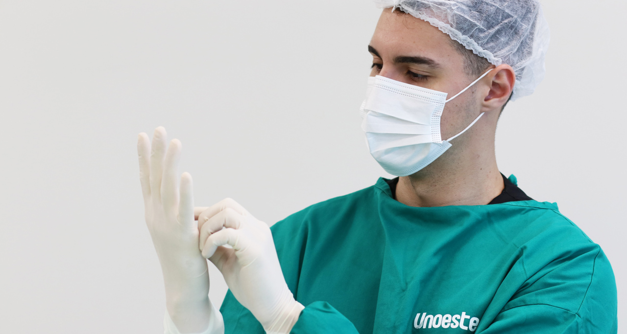 Estudante de Medicina da Unoeste vestindo avental cirúrgico verde, máscara e touca descartável, colocando luvas estéreis antes de iniciar atividade prática em ambiente hospitalar.