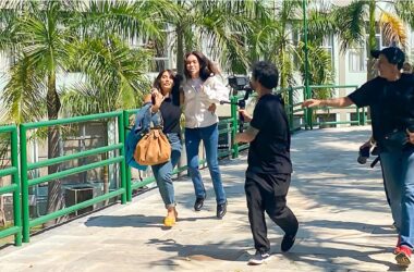 Duas mulheres correm lado a lado em uma passarela elevada com grades verdes, enquanto um homem com uma câmera as segue de perto, e outro homem, possivelmente da equipe de filmagem, os acompanha. Ao fundo, palmeiras e um edifício claro podem ser vistos. A imagem captura um momento de gravação do filme “Deixe-me Viver” na Unoeste.