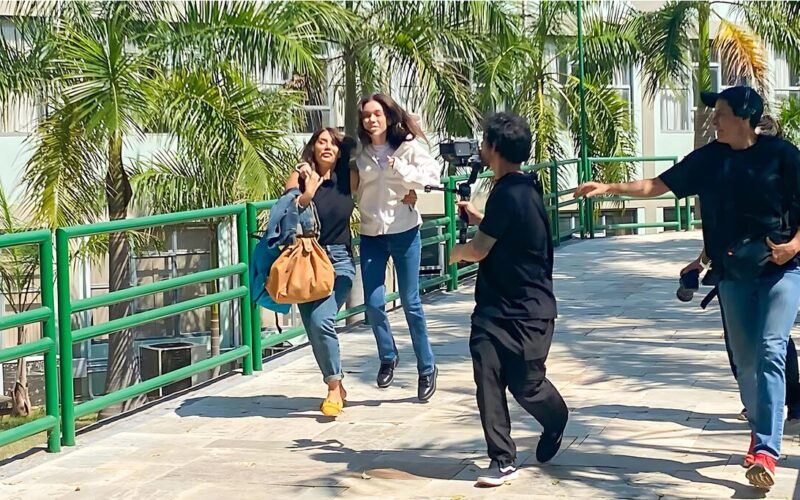 Duas mulheres correm lado a lado em uma passarela elevada com grades verdes, enquanto um homem com uma câmera as segue de perto, e outro homem, possivelmente da equipe de filmagem, os acompanha. Ao fundo, palmeiras e um edifício claro podem ser vistos. A imagem captura um momento de gravação do filme “Deixe-me Viver” na Unoeste.