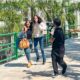 Duas mulheres correm lado a lado em uma passarela elevada com grades verdes, enquanto um homem com uma câmera as segue de perto, e outro homem, possivelmente da equipe de filmagem, os acompanha. Ao fundo, palmeiras e um edifício claro podem ser vistos. A imagem captura um momento de gravação do filme “Deixe-me Viver” na Unoeste.