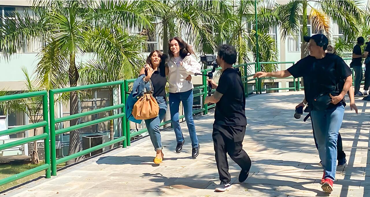 Duas mulheres correm lado a lado em uma passarela elevada com grades verdes, enquanto um homem com uma câmera as segue de perto, e outro homem, possivelmente da equipe de filmagem, os acompanha. Ao fundo, palmeiras e um edifício claro podem ser vistos. A imagem captura um momento de gravação do filme “Deixe-me Viver” na Unoeste.
