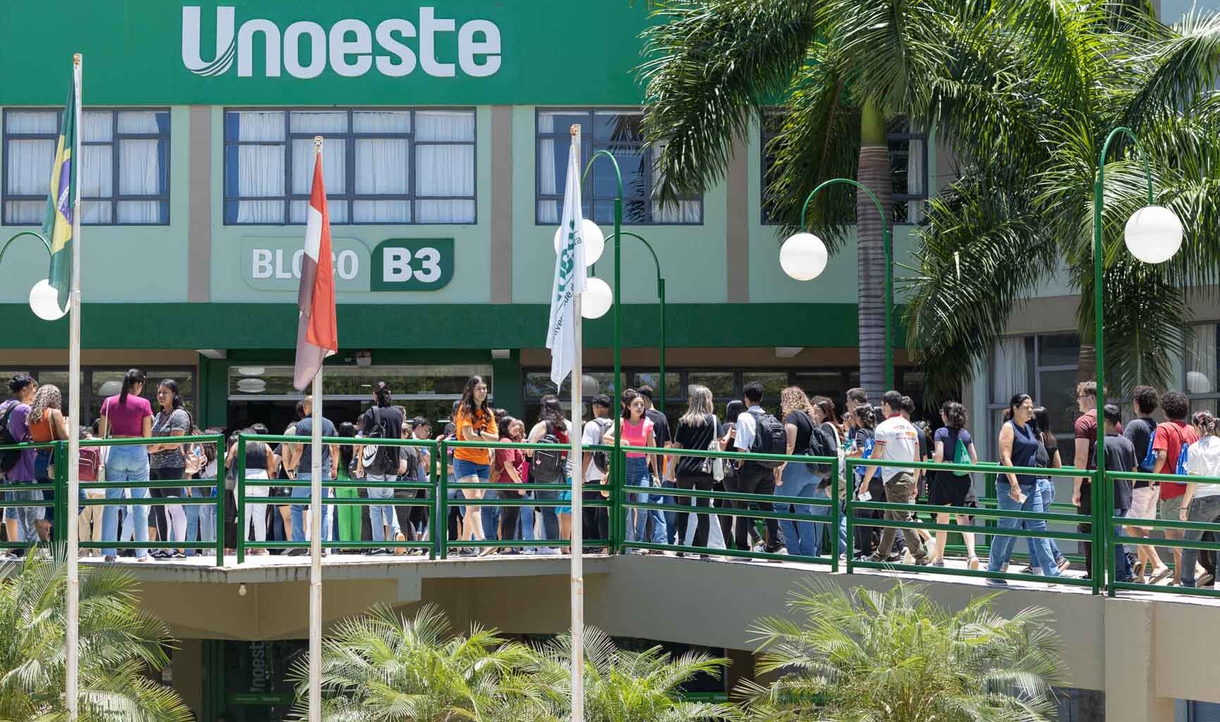 Dia de ENEM na Unoeste: Estudantes caminham em passarela verde em frente ao Bloco B3 da universidade, com palmeiras e postes de luz ao redor.