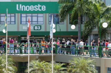 Dia de ENEM na Unoeste: Estudantes caminham em passarela verde em frente ao Bloco B3 da universidade, com palmeiras e postes de luz ao redor.