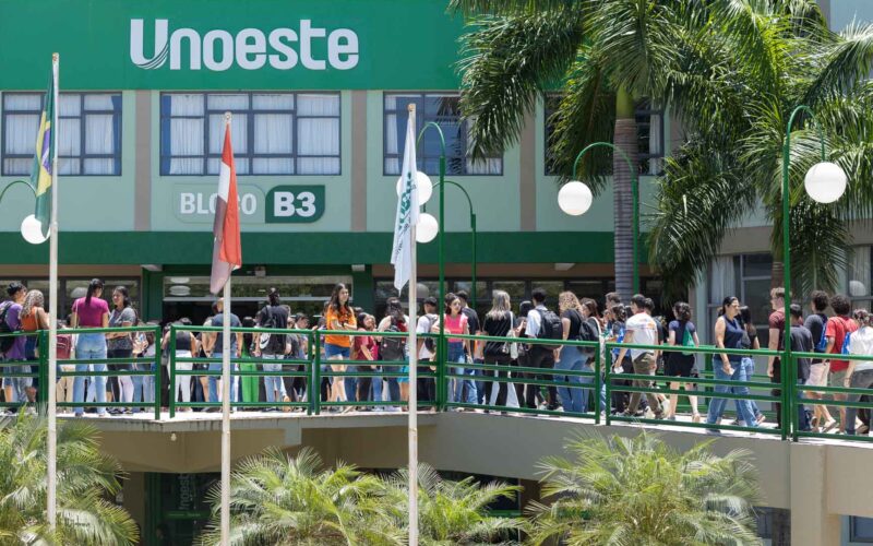 Dia de ENEM na Unoeste: Estudantes caminham em passarela verde em frente ao Bloco B3 da universidade, com palmeiras e postes de luz ao redor.