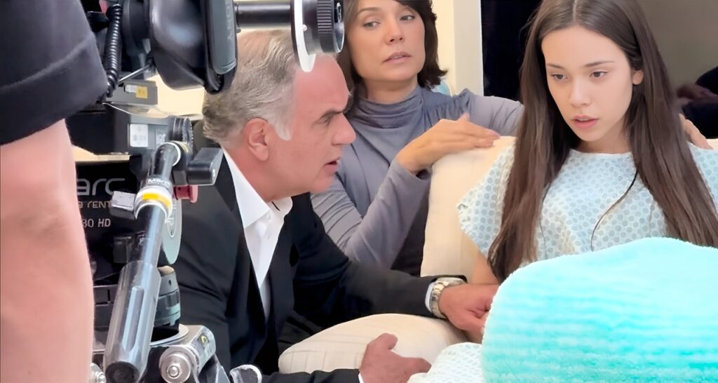 Julia, uma jovem de cabelos castanhos e expressão preocupada, vestindo uma bata hospitalar azul clara, está sentada em uma cama, com seu pai, um homem de cabelos grisalhos e terno escuro segurando sua mão e olhando para ela com seriedade. Atrás deles, a mão, mulher de cabelos escuros e blusa cinza, observa a cena com uma expressão pensativa. Um equipamento de câmera pode ser visto no canto inferior esquerdo, sugerindo que a cena está sendo filmada.