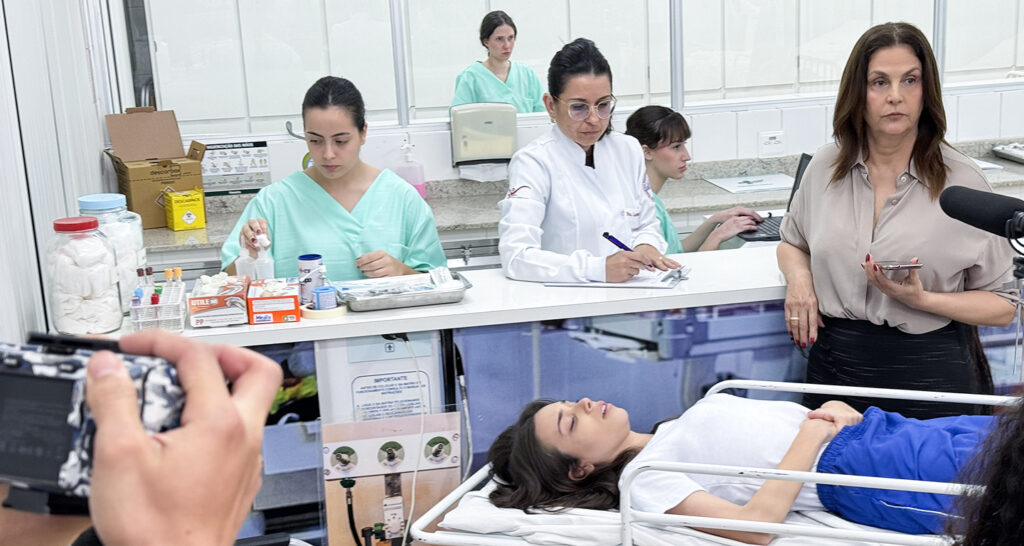 Julia, jovem de cabelos castanhos, vestindo uma camiseta branca e shorts azuis, está deitada em uma maca de hospital com os olhos fechados. Ao redor dela, em um ambiente médico, várias pessoas estão trabalhando. Duas mulheres de jaleco branco e óculos estão em uma bancada, uma escrevendo e a outra manuseando materiais. Outras duas mulheres de uniforme verde claro estão ao fundo. No primeiro plano, uma mão segura uma câmera, indicando que a cena está sendo filmada.