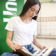 Jovem estudante sentada no campus da Unoeste, usando óculos e camiseta branca, lendo um livro aberto sobre livros que viraram filmes, ao lado de um totem verde com o logo da universidade, em ambiente interno com piso claro e paredes revestidas com azulejos.