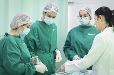 Estudantes de Enfermagem da Unoeste, com jalecos e máscaras, recebem orientação prática de docente em ambiente de laboratório.