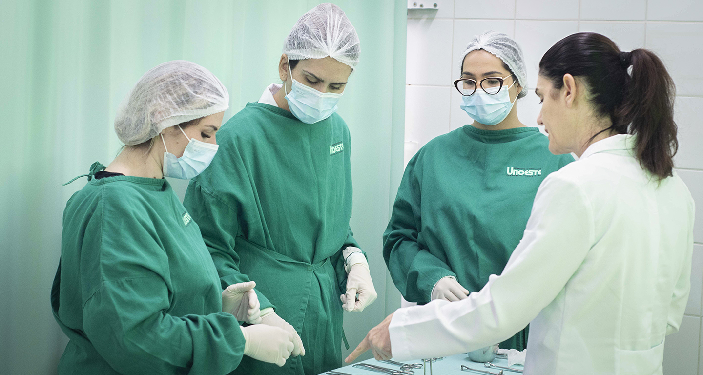 Estudantes de Enfermagem da Unoeste, com jalecos e máscaras, recebem orientação prática de docente em ambiente de laboratório.