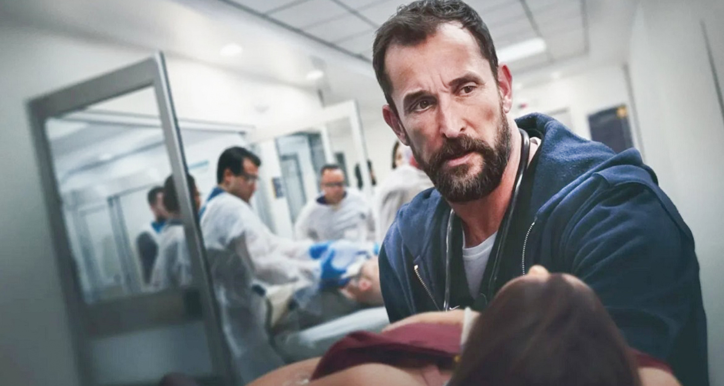 Dr. Michael encara paciente em estado crítico enquanto equipe atua ao fundo, cena intensa da série médica The Pitt da HBO.