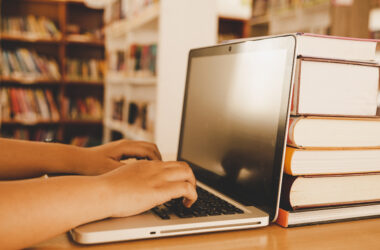 Estudante digitando em um notebook sobre uma mesa de biblioteca, com pilha de livros ao lado e prateleiras ao fundo