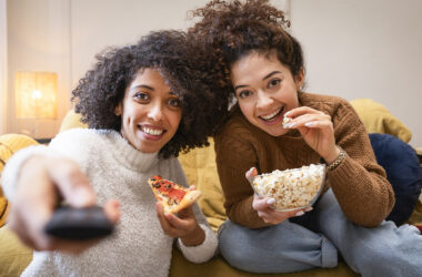 Duas jovens assistindo a filmes de animação no sofá, uma comendo pizza e segurando controle remoto e a outra saboreando pipoca em um ambiente aconchegante.