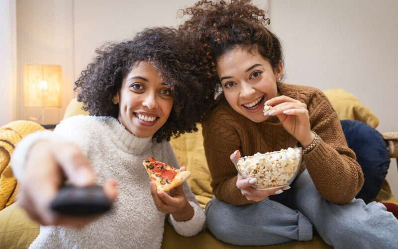 Duas jovens assistindo a filmes de animação no sofá, uma comendo pizza e segurando controle remoto e a outra saboreando pipoca em um ambiente aconchegante.