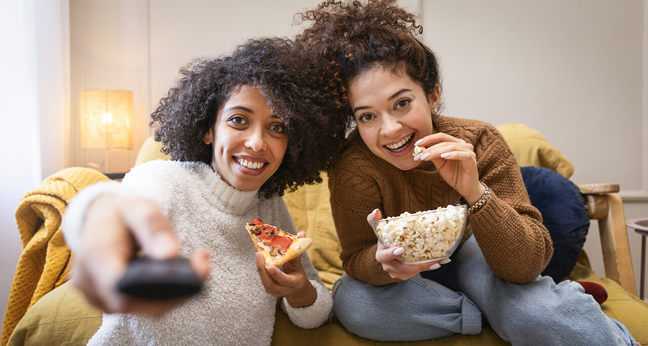 Duas jovens assistindo a filmes de animação no sofá, uma comendo pizza e segurando controle remoto e a outra saboreando pipoca em um ambiente aconchegante.