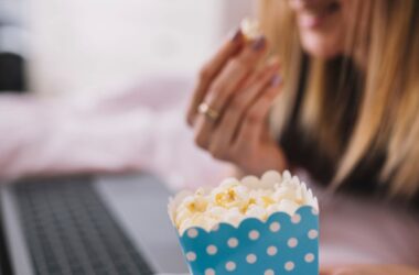 Mulher assistindo filme em um notebook enquanto segura pipoca em embalagem azul com bolinhas brancas, parcialmente desfocada ao fundo.