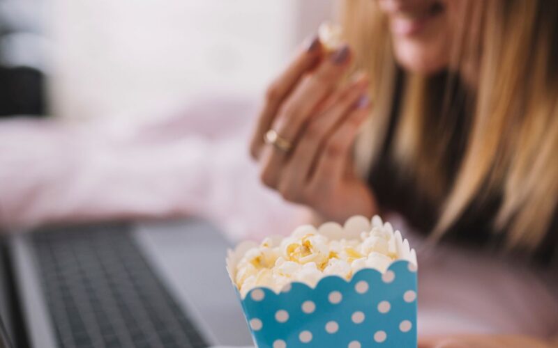 Mulher assistindo filme em um notebook enquanto segura pipoca em embalagem azul com bolinhas brancas, parcialmente desfocada ao fundo.