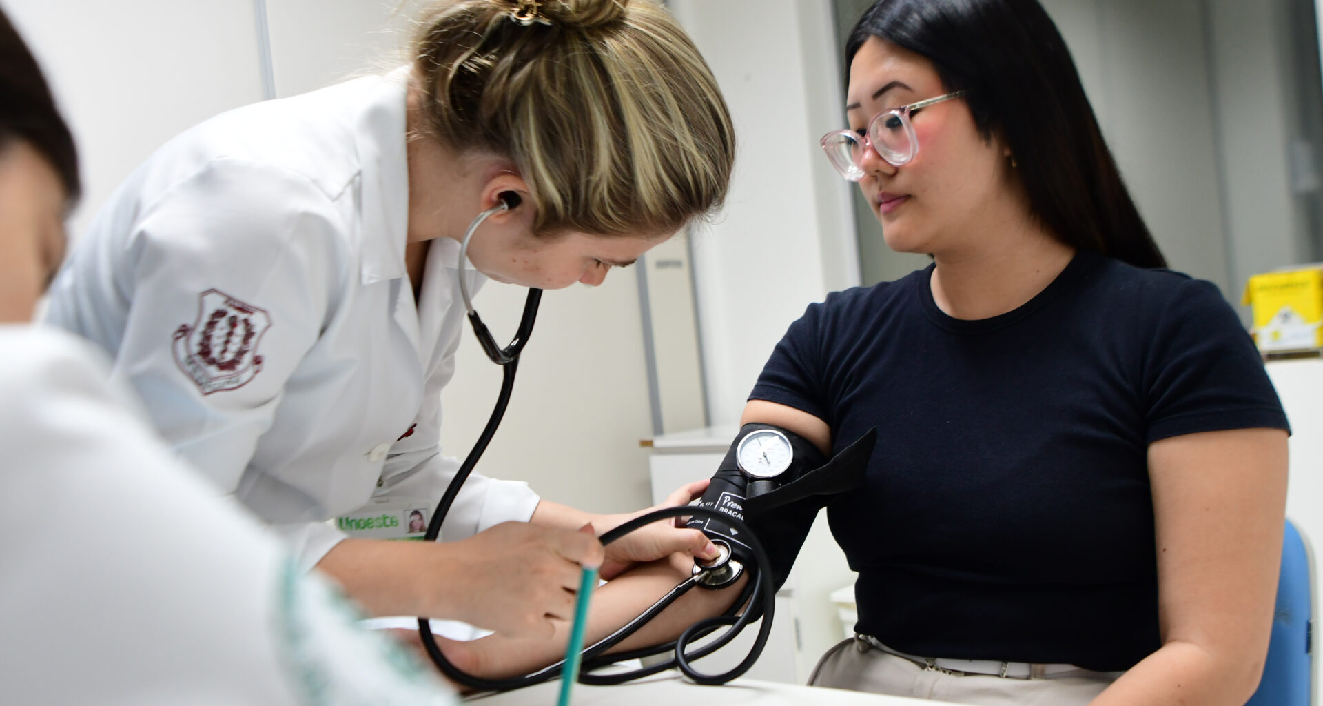 Estudante de Medicina da Unoeste mede a pressão arterial de uma paciente durante atendimento clínico, utilizando estetoscópio e esfigmomanômetro em ambiente acadêmico.