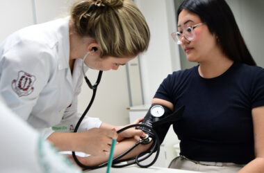 Estudante de Medicina da Unoeste mede a pressão arterial de uma paciente durante atendimento clínico, utilizando estetoscópio e esfigmomanômetro em ambiente acadêmico.