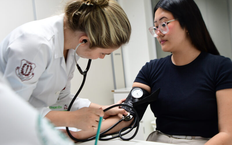 Estudante de Medicina da Unoeste mede a pressão arterial de uma paciente durante atendimento clínico, utilizando estetoscópio e esfigmomanômetro em ambiente acadêmico.