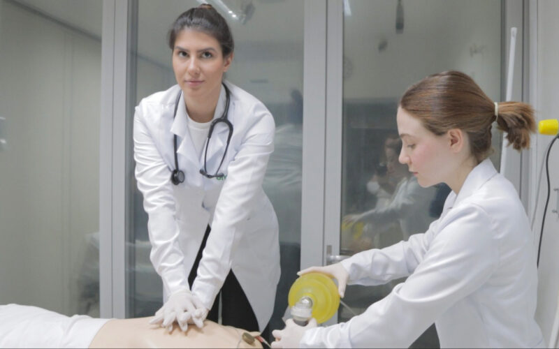 Duas estudantes de Medicina da Unoeste realizam treinamento prático em laboratório: uma executa massagem cardíaca em manequim de simulação, enquanto a outra auxilia com ventilação manual, representando a atuação das mulheres na Medicina.
