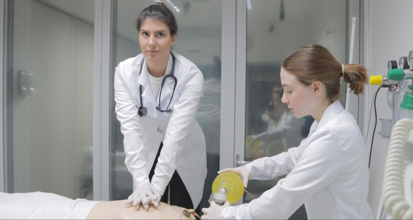 Duas estudantes de Medicina da Unoeste realizam treinamento prático em laboratório: uma executa massagem cardíaca em manequim de simulação, enquanto a outra auxilia com ventilação manual, representando a atuação das mulheres na Medicina.